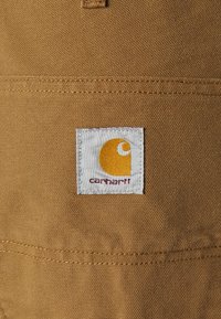 Carhartt logo patch on a tan fabric with a textured surface; patch features a yellow and white design with text below.