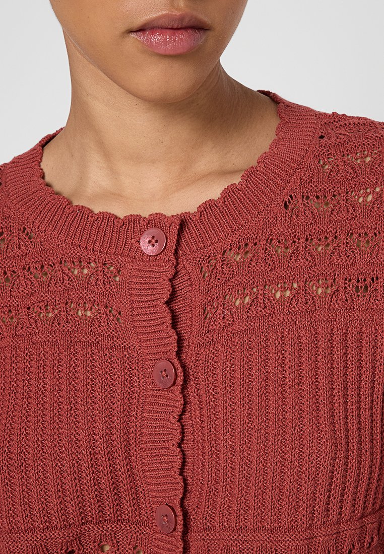Nahaufnahme einer Person, die eine strukturierte, rostfarbene Strickjacke mit gewellten Kanten und drei passenden Knöpfen trägt.