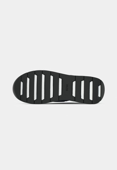Semelle de chaussure noire avec un design en caoutchouc texturé, présentant de larges rainures, des accents blancs et une marque sur la surface.