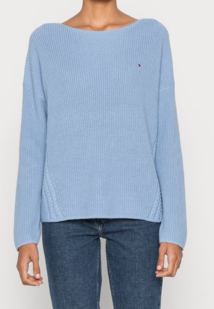 Persoon draagt een lichtblauwe gebreide trui met lange mouwen en donkerblauwe spijkerbroek, staand tegen een effen lichte achtergrond.