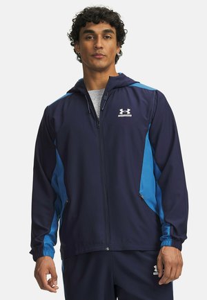 Mann trägt eine marineblaue und blaue Under Armour Zip-Hoodie-Jacke und passende Hose vor einem einfarbigen Hintergrund.