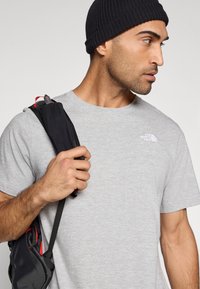 Γκρι βαμβακερό μπλουζάκι με κοντά μανίκια και στρογγυλή λαιμόκοψη. Μαύρο σκούφο και μαύρη τσάντα με κόκκινες λεπτομέρειες που κρατάει το πρόσωπο.