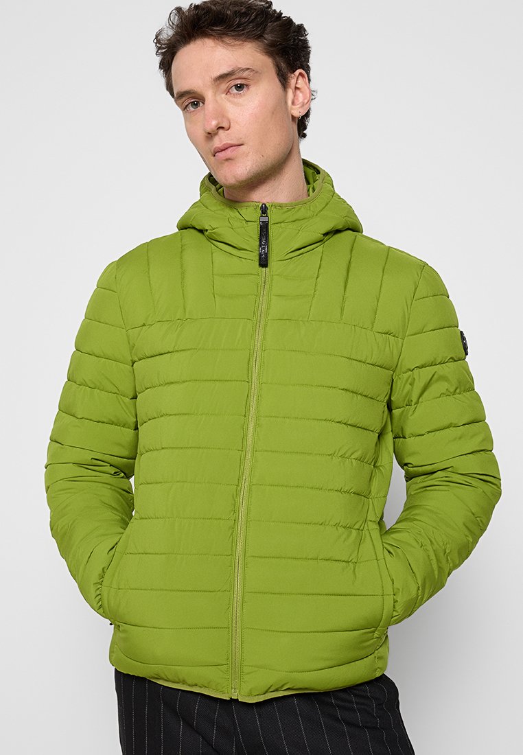 Uomo che indossa una giacca con cappuccio trapuntata verde lime con zip, mani nelle tasche, e pantaloni neri a righe sottili, contro uno sfondo semplice.