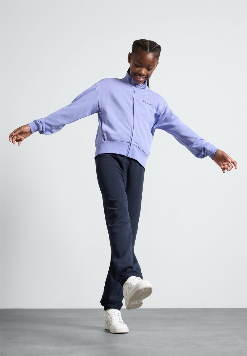 Giacca zip-up lavanda con colletto alto e orlo a costine, abbinata a pantaloni sportivi blu navy e sneakers bianche su una superficie grigia.