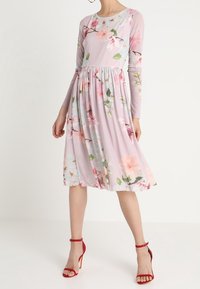 Pitkähihainen, liila mekko, jossa on pinkki- ja vihervärinen kukkakuvio. Flareskirtissä on poimut, ja sen kanssa on yhdistetty punaiset remmisandaalit.