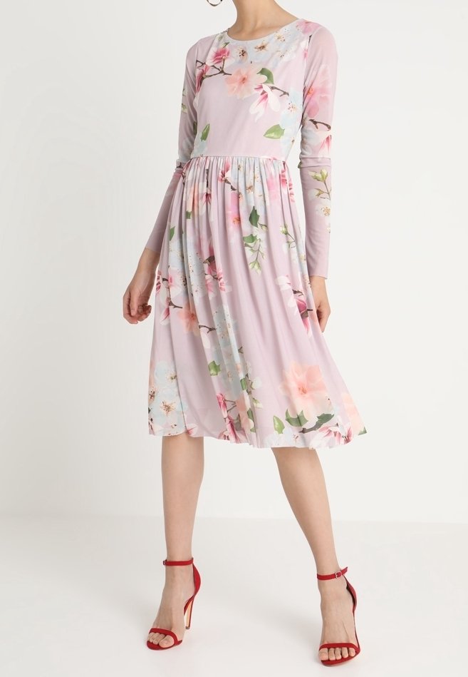 Pitkähihainen, liila mekko, jossa on pinkki- ja vihervärinen kukkakuvio. Flareskirtissä on poimut, ja sen kanssa on yhdistetty punaiset remmisandaalit.