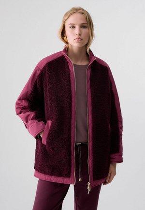 Young woman wearing a dark purple fleece jacket with quilted sleeves and pants against a plain light background.