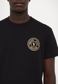 Tricou din bumbac negru cu un logo circular aurit care conține "VERSACE JEANS COUTURE" și o literă centrală "V" pe pieptul stâng.