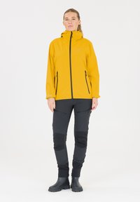 Gul vandtæt jakke med sorte lynlåse, parret med sorte bukser med lynlåslommer og knælapper. Sorte gummistøvler fuldender outfittet.