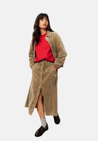 Jupe maxi en velours côtelé de couleur marron clair avec des boutons à l'avant et des poches latérales, associée à un pull rouge et des chaussures noires. Le modèle se tient les mains dans les poches.