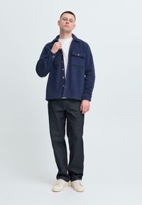 Camisa chaqueta de forro polar azul marino con cierre de botones, dos bolsillos frontales y corte relajado, combinada con pantalones de mezclilla oscuros y zapatillas beige.