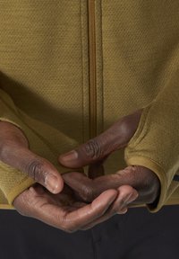 Długi rękaw, zapinany na zamek top w musztardowym kolorze, z teksturowanej tkaniny, z otworami na kciuki w mankietach oraz pół zamkiem z przodu. Ręce złożone razem.