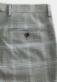 Grau karierte Hose mit strukturierter Oberfläche. Besitzt eine Gesäßtasche mit einem schwarzen Knopf und einen Bund mit Gürtelschlaufen.