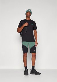 Ung mann med lue, svart t-skjorte, grønne og svarte shorts og joggesko, stående med ryggsekkstropp over skulderen mot ensfarget bakgrunn.