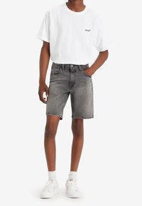 Bermudas de mezclilla gris con un corte clásico, bolsillos frontales y una línea de cinturón visible, combinadas con una camiseta blanca lisa que presenta un pequeño logo.