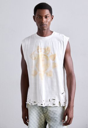 Ärmelloses weißes T-Shirt mit einem ausfransenden Saum, das ein verblasstes gelbes Sonnenmotiv zeigt. Kombiniert mit hellblauen gemusterten Jeans.