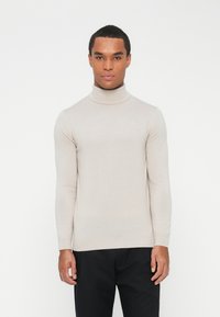 Bež turtleneck pulover, usklađen dizajn, duge rukave, fina tekstura, suptilan logotip na prsima, nosi se s tamnim hlačama.