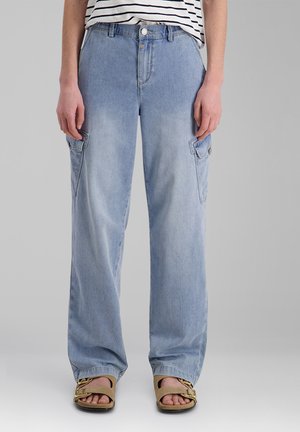 Person trägt hellblaue, locker sitzende Cargo-Jeans mit Seitentaschen, ein gestreiftes Hemd und beige Sandalen mit goldenen Kettendetails.