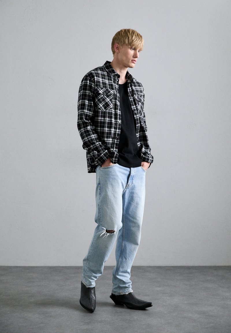 Chemise en flanelle à carreaux noir et blanc portée sur un t-shirt noir, associée à un jean bleu clair déchiré et à des bottes en cuir noires à bouts pointus.