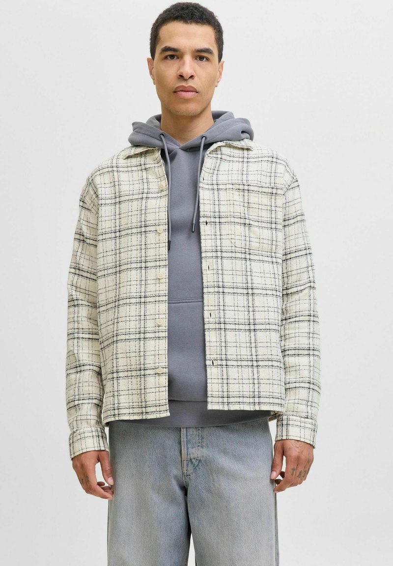 Beige und schwarze karierte Flanelle-shirt mit Knöpfen, über einem grauen Hoodie getragen, kombiniert mit hellblauen Jeans.