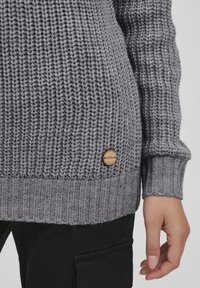Grauer Strickpullover mit gerippten Bündchen und Saum. Verfügt über ein kleines Holzetikett mit "OXMO" an der Seite. Die Textur wirkt dick und gemütlich.