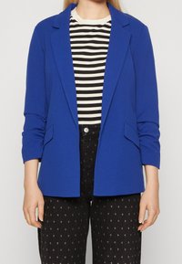 Modrá blazer se zakázkovým střihem, s jednobuttonovým zapínáním, dvěma předními kapsami a nařasenými rukávy, nošená přes pruhované tričko.