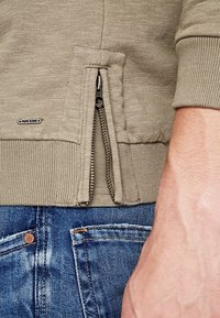 Felpa beige con zip, in tessuto texture, dotata di tasca laterale con zip e polsini a costine, abbinata a jeans in denim blu.