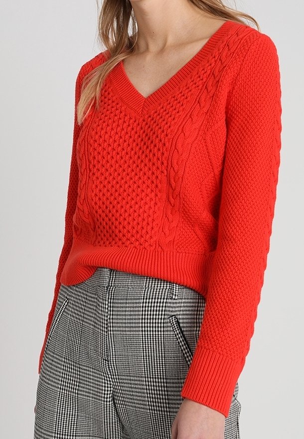 Pull tricoté rouge à col en V, avec des motifs texturés et des poignets côtelés. Associé à un pantalon à carreaux noir et blanc.