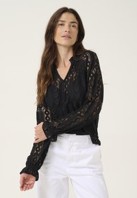 Black lace blouse with a V-neck, featuring intricate patterns, long sleeves, and a ruffled hem detail. Styled with white trousers.