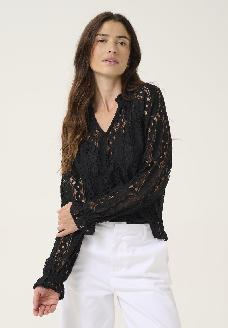 Black lace blouse with a V-neck, featuring intricate patterns, long sleeves, and a ruffled hem detail. Styled with white trousers.