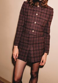 Brunt ternet dragt sæt med en figursyet jakke med guldknapper, lange ærmer og en plisseret mininederdel, tekstureret med metalliske tråde.