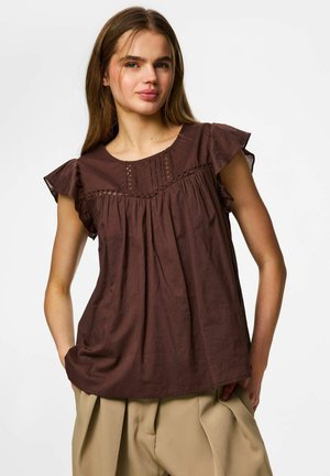 Jeune femme aux longs cheveux bruns portant un haut marron ample sans manches et un pantalon beige plissé, debout les mains dans les poches.