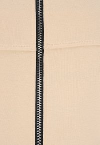 Beige szövet, texturált felülettel, középen fekete cipzárral és kontrasztos ezüst villákkal. Egyenes vonalú szabás, tiszta varratokkal.