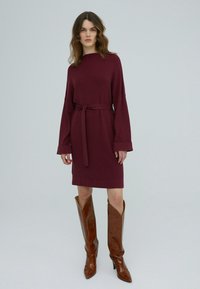 Burgunderfarbene Strickkleid mit einem Gürtel, langen Ärmeln und Manschettenakzenten, kombiniert mit hohen braunen Lederstiefeln mit spitzen Zehen und strukturiertem Finish.