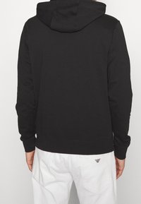 Persona con una sudadera con capucha negra y pantalones blancos con un pequeño logo en el bolsillo trasero, de pie contra un fondo liso.