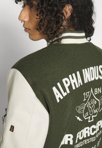 Giacca varsity verde in misto lana con maniche bianche. Presenta testo e grafiche ricamate sul retro. Collo a costine con dettagli a strisce.