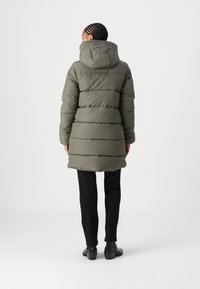 Manteau rembourré vert olive, jusqu'aux genoux, avec capuche, présentant des motifs matelassés horizontaux et une finition texturée, associé à un pantalon noir et des bottines.