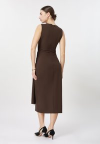 Femme de dos portant une robe midi marron sans manches et ajustée avec une fermeture éclair dans le dos, et des chaussures à talons hauts noires, debout sur un fond uni.