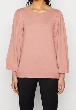 Kvinde iført en støvet lyserød langærmet sweater med puffede ærmer og sorte bukser, stående mod en ensfarvet lys baggrund.