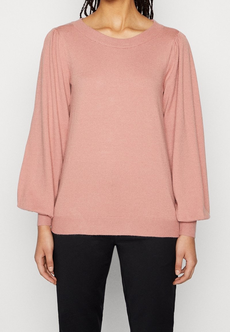 Femme portant un pull rose poussiéreux à manches longues bouffantes et un pantalon noir, debout devant un fond clair uni.