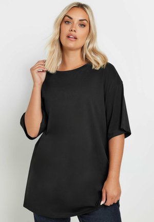 Blondynka o włosach do ramion, ubrana w luźną czarną koszulkę z krótkim rękawem, na jednolitym jasnym tle.