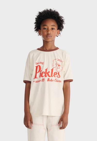 Bambino con capelli ricci che indossa una maglietta beige con rifiniture marroni, testo rosso "Pickles" e grafica di un cane, abbinata a pantaloni chiari.