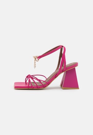 Roze sandalen met bandjes en een vierkante neus, verstelbare enkelband met gouden ketting en brede trapeziumvormige blokhak op een witte achtergrond.
