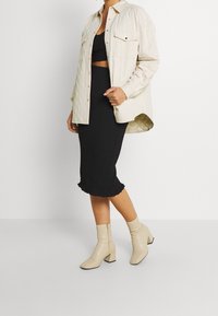 Camisa chaqueta acolchada beige sobre un top corto negro de canalé y una falda negra hasta la rodilla con dobladillo con volantes. Botines de tacón cuadrado beige.
