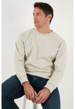 Vaalea beige collegepaita, valmistettu pehmeästä kankaasta, jossa pyöreä pääntie, pitkät raglanhihat ja rintapielessä pieni logo. Yhdistetty tummien farkkujen kanssa.