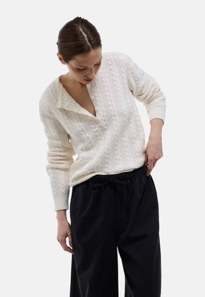 Femme portant un pull en maille torsadée écru avec un large col et un pantalon noir ample, regardant vers le bas et ajustant sa ceinture.