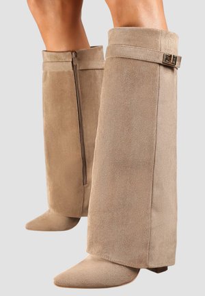 Bottes hauteur genou en suède beige avec fermeture éclair latérale sur une botte et sangle décorative avec boucle sur l'autre, portées sur des jambes nues.