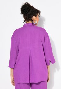 Blusa viola oversize con colletto, maniche a tre quarti e dettaglio di piega sul retro. Realizzata in tessuto testurizzato con una leggera lucentezza.