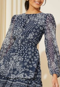 Navy blue dress with white floral and geometric patterns, sheer puff sleeves, a fitted waist, and a tiered skirt design.