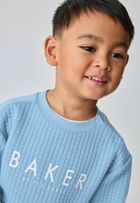 Lys blå textured sweatshirt med et ribbet mønster, prydet med hvidt "BAKER" logo. Trykknapper på skulderen tilføjer detaljer.